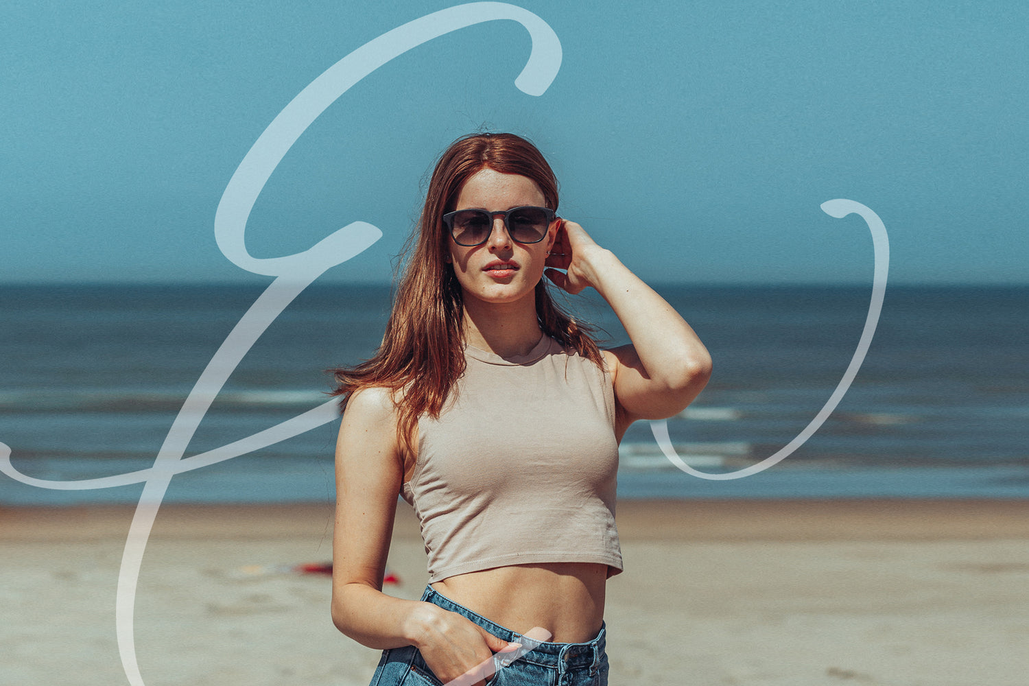 Er staat een mevrouw op het strand met een zwarte zonnebril. Ze gaat met haar hand door haar haar en in de achtergrond is een logo te zien van EveryWear.