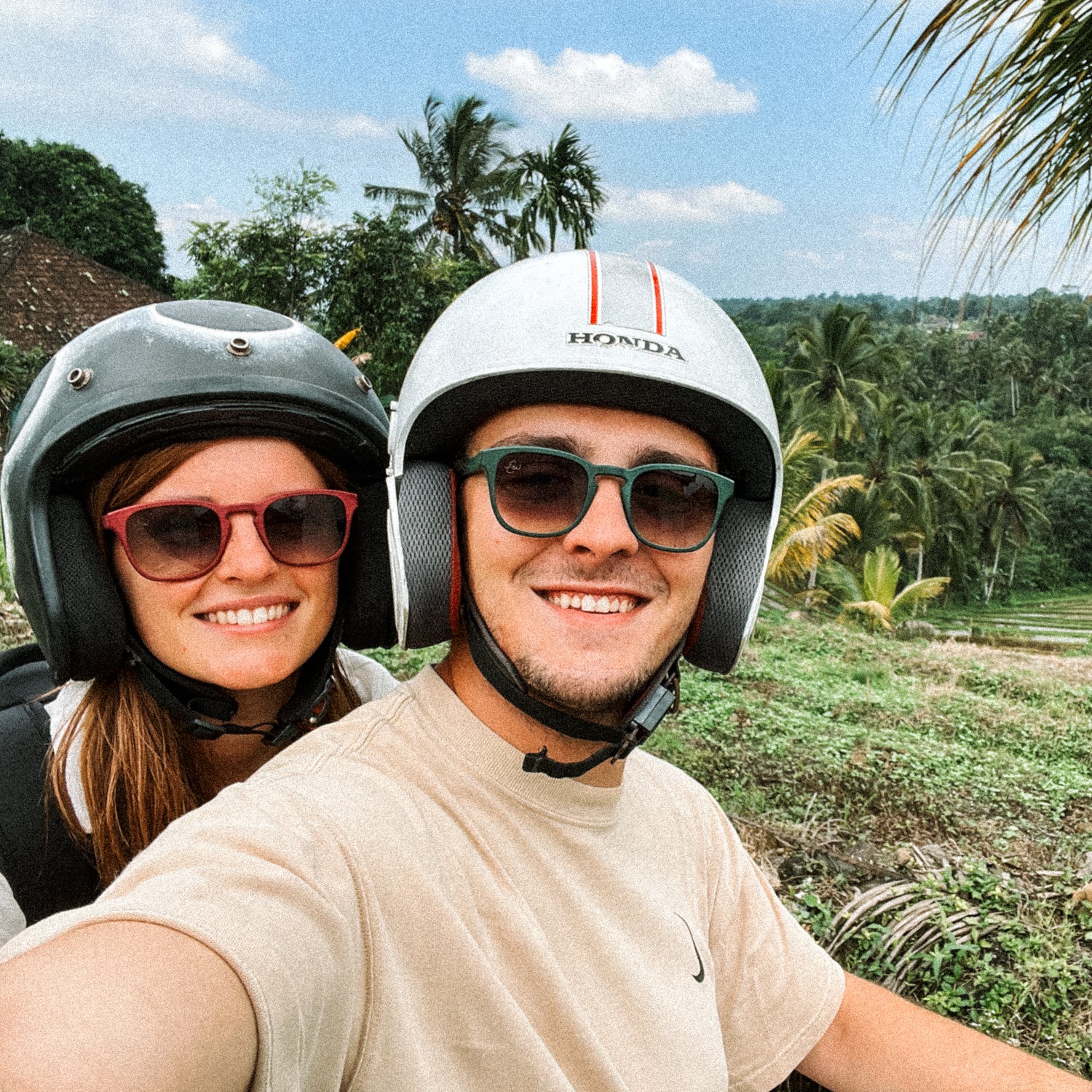 Twee personen, die waarschijnlijk op een scooter zitten, maken een selfie op Bali. Beide personen hebben een zonnebril op, de een een rode, de ander een groene.