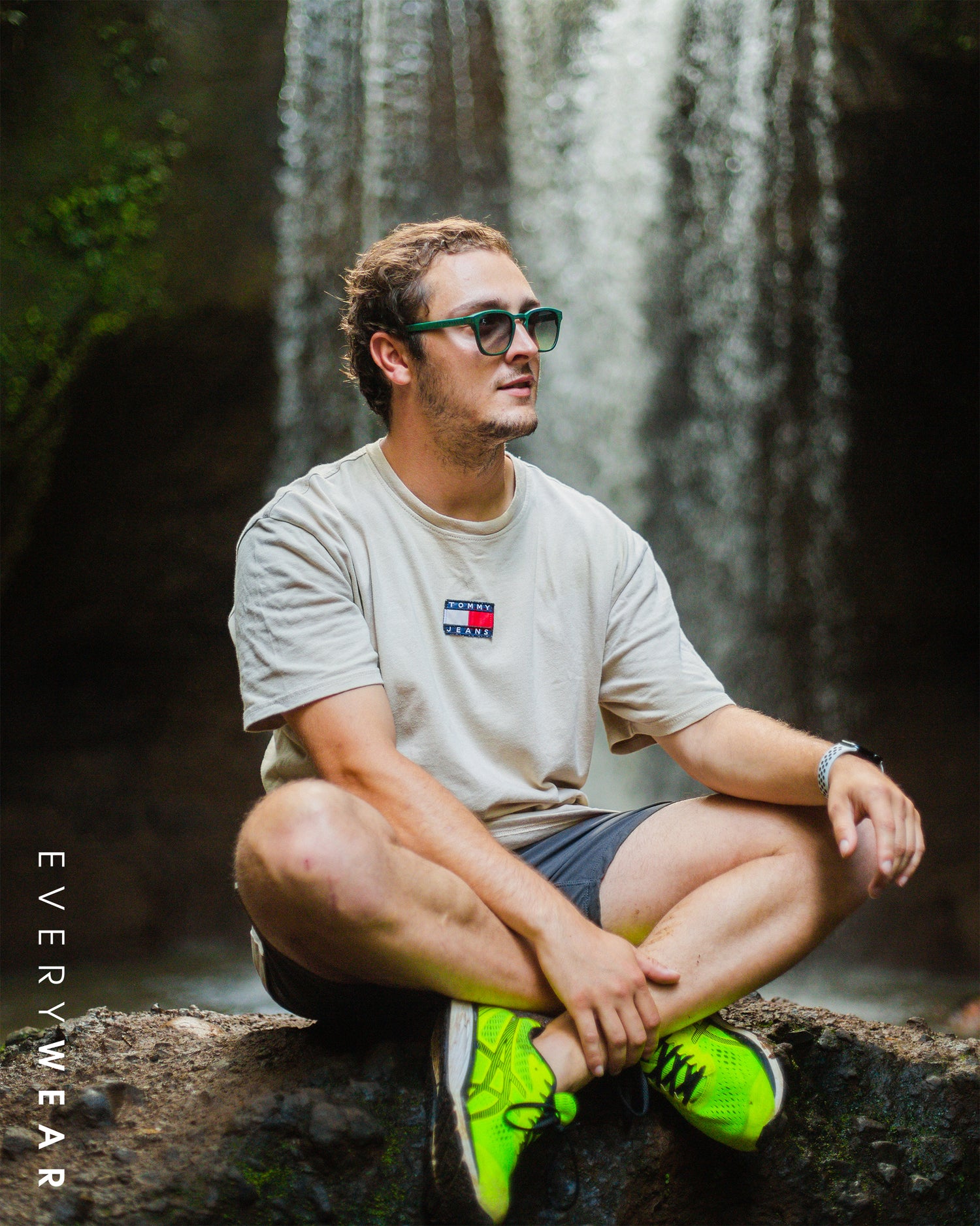 Portretfoto van een man zittend op een rots in kleermakerszit. Op de achtergrond is een waterval te zien en hij draagt een groene zonnebril.
