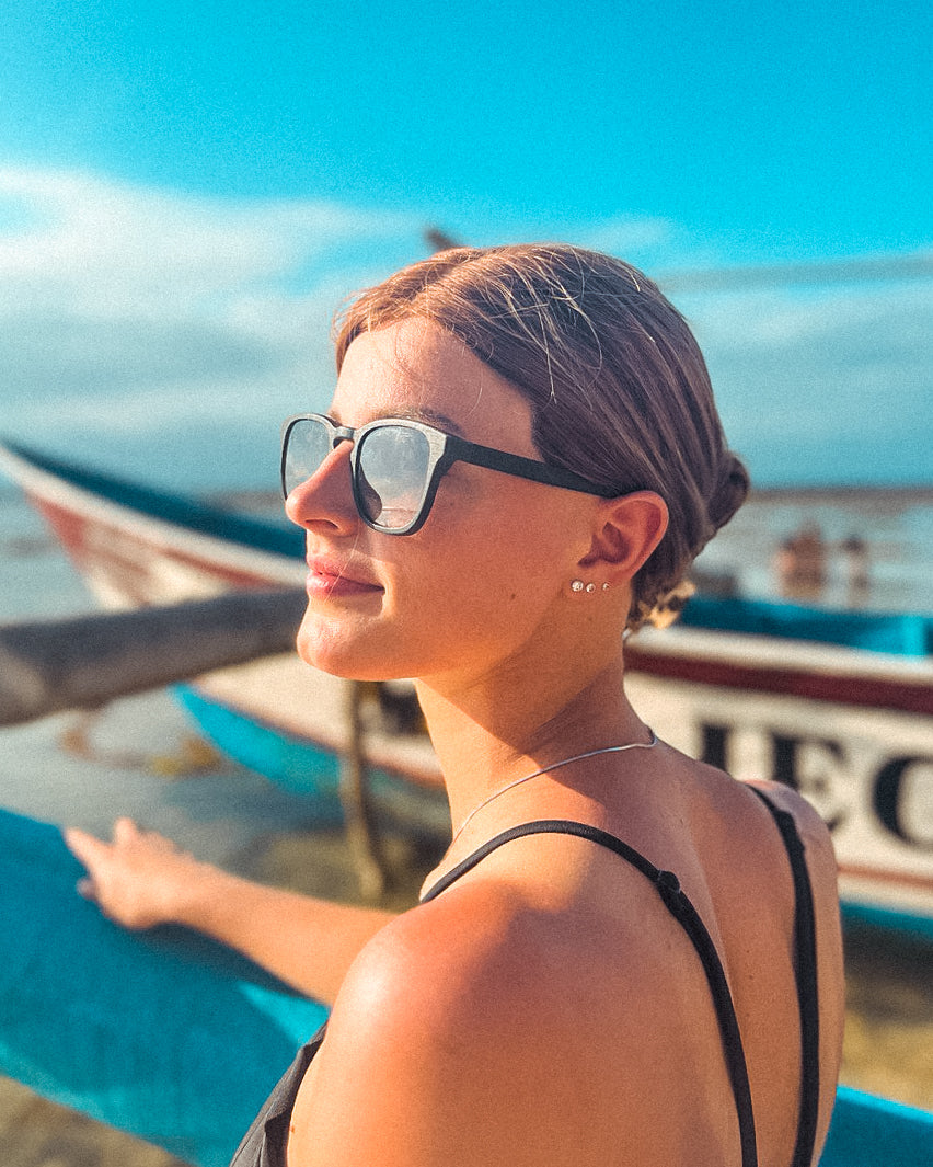 Zwarte zonnebril gedragen door een vrouw. In de wazige achtergrond is een gedeelte van een boot te zien en een strand.
